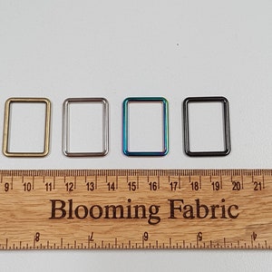 Op de afbeelding: Vier rechthoekige metalen ringen in verschillende afwerkingen: goud, zilver, iriserend en zwart. De ringen zijn uitgelijnd op een houten liniaal met de tekst "Blooming Fabric" erop gedrukt.