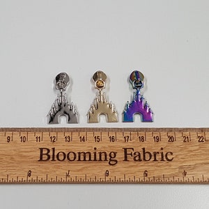 May include: Three zipper pulls in the shape of a castle, one silver, one gold, and one iridescent. The zipper pulls are lined up on a wooden ruler with the numbers 7 through 24 visible.