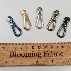 May include: Five zipper pulls in different colours: iridescent, gold, silver, brass, and black. The zipper pulls are all the same size and shape, with a small loop at the top and a larger loop at the bottom. The zipper pulls are arranged in a row on a wooden ruler with the text "Blooming Fabric" printed on it.