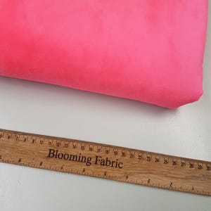 K&ouml;nnte beinhalten: Nahaufnahme eines leuchtend pinkfarbenen Stoffmusters. Ein Holzliniall mit der Aufschrift "Blooming Fabric" ist im unteren Bildbereich sichtbar.