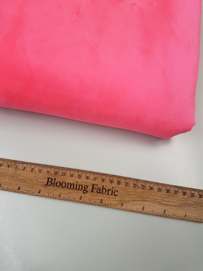 K&ouml;nnte beinhalten: Nahaufnahme eines leuchtend pinkfarbenen Stoffmusters. Ein Holzliniall mit der Aufschrift "Blooming Fabric" ist im unteren Bildbereich sichtbar.