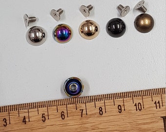 May include: Assortment of seven metal dome-shaped studs with screws. The studs are silver, gold, black, iridescent, and bronze. A ruler is in the background.
