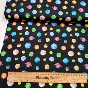 May include: Black fabric with a repeating pattern of colorful planets and stars. The fabric is shown with a ruler that reads "Blooming Fabric".