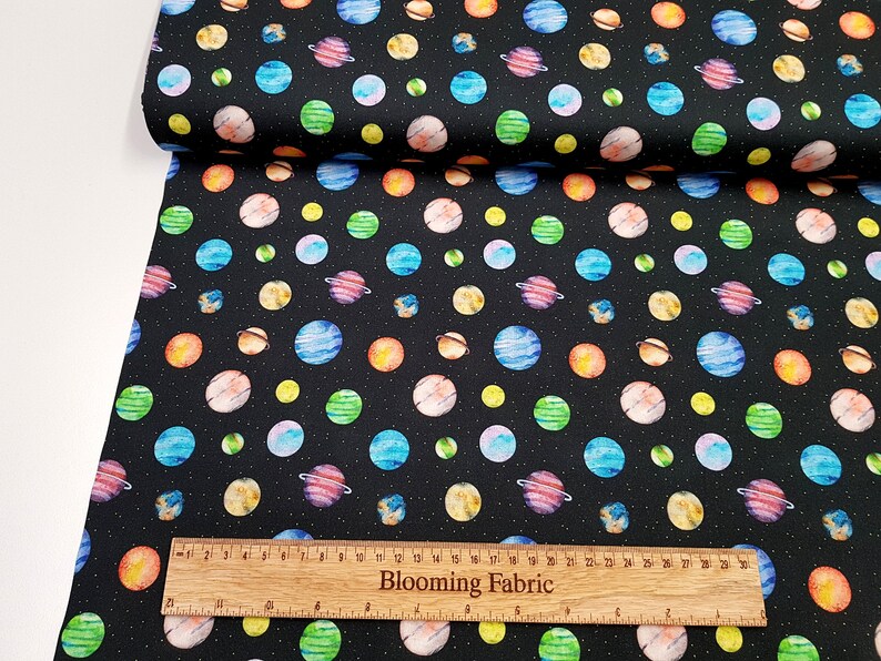 May include: Black fabric with a repeating pattern of colorful planets and stars. The fabric is shown with a ruler that reads "Blooming Fabric".