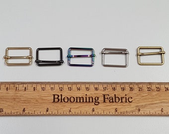 May include: Five metal slider buckles in different finishes: gold, black, iridescent, silver, and brass. The buckles are lined up on a wooden ruler with the numbers 5 through 24 visible.