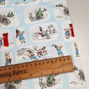 Può includere: Tessuto blu e bianco con un motivo ripetuto di personaggi di Peter Rabbit in varie scene invernali. Le scene includono Peter Rabbit che consegna una calza, Peter Rabbit che consegna la posta, Peter Rabbit che pattina sul ghiaccio e Peter Rabbit che tira un albero di Natale.