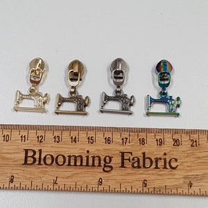 Puede incluir: Cuatro tiradores de cremallera con un diseño de máquina de coser. Los tiradores son de color dorado, dorado antiguo, plateado e iridiscente. Los tiradores se muestran en una regla de madera con el texto "Blooming Fabric".