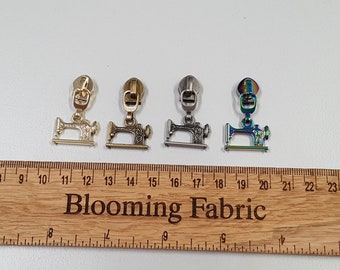 Pode incluir: Quatro puxadores de zíper com um design de máquina de costura. Os puxadores são nas cores dourado, dourado antigo, prata e iridescente. Os puxadores são mostrados em uma régua de madeira com o texto "Blooming Fabric".