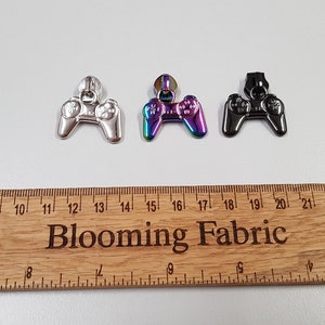 Peut inclure: Trois tirettes de fermeture éclair en métal de couleurs différentes en forme de manettes de jeu vidéo. Les tirettes sont argentées, irisées et noires.