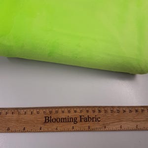 K&ouml;nnte beinhalten: Nahaufnahme eines leuchtend gr&uuml;nen Stoffmusters. Der Stoff ist weich und pl&uuml;schig. Ein Holzliniall mit der Aufschrift "Blooming Fabric" ist am unteren Rand des Bildes zu sehen.