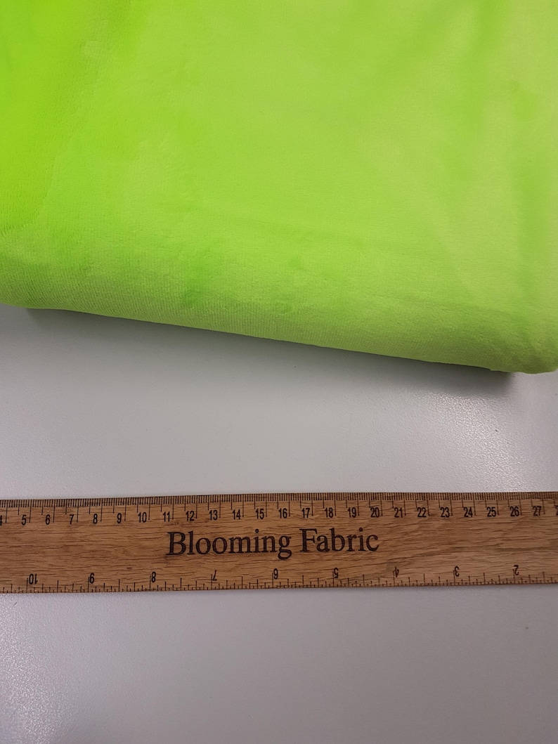 K&ouml;nnte beinhalten: Nahaufnahme eines leuchtend gr&uuml;nen Stoffmusters. Der Stoff ist weich und pl&uuml;schig. Ein Holzliniall mit der Aufschrift "Blooming Fabric" ist am unteren Rand des Bildes zu sehen.