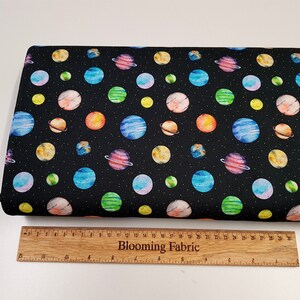 May include: Black fabric with a repeating pattern of colorful watercolor planets and stars. The fabric is shown next to a wooden ruler with the text "Blooming Fabric" printed on it.