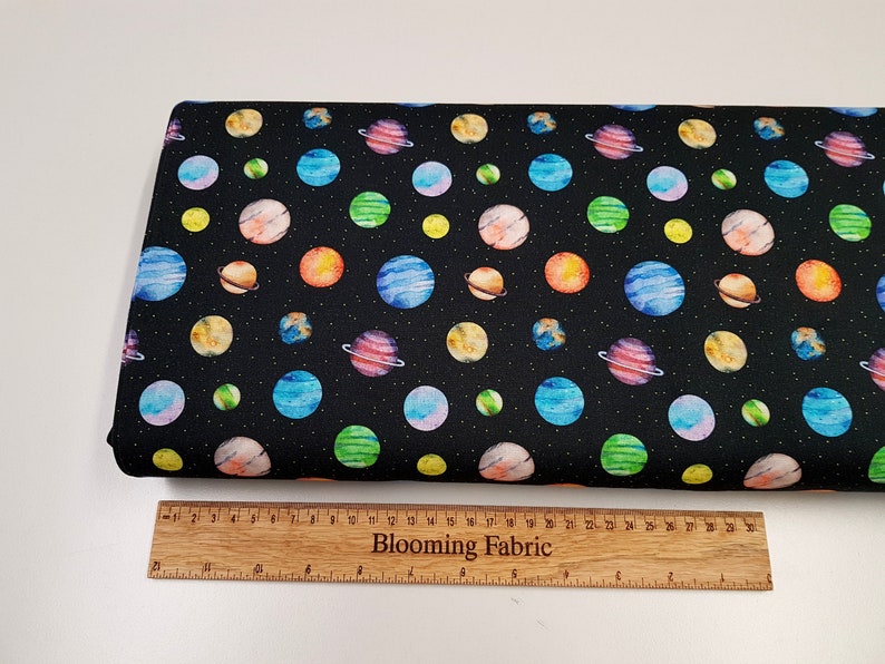 May include: Black fabric with a repeating pattern of colorful watercolor planets and stars. The fabric is shown next to a wooden ruler with the text "Blooming Fabric" printed on it.