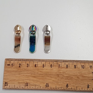Puede incluir: Se muestran tres tiradores de cremallera metálicos en tonos dorados, iridiscentes y plateados. Una regla de madera con marcas en pulgadas y centímetros se coloca junto a los tiradores. Los tiradores de cremallera miden aproximadamente 2,5 cm de largo.