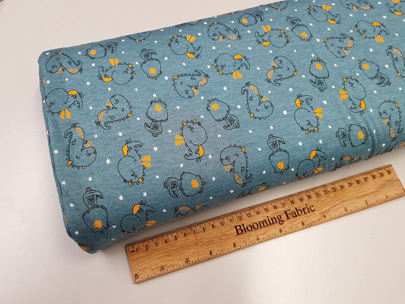 May include: Blue fabric with a white star pattern and a repeating pattern of yellow and black cartoon dragons. The fabric is measured with a wooden ruler that says "Blooming Fabric".