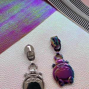 Może przedstawiać: Dwa srebrne suwaki do zamka błyskawicznego z wzorem Totoro. Jeden suwak jest srebrny, a drugi tęczowy.