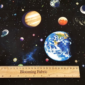 Puede incluir: Tela negra con un estampado de temática espacial que presenta planetas, estrellas y una galaxia. La tela incluye una regla con el texto "Blooming Fabric" impreso en ella.