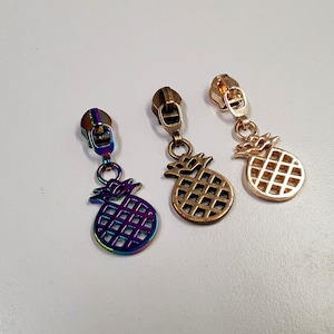 Op de afbeelding: Drie ritsen met ananas-bedels. De bedels zijn goud, regenboogkleurig en zilver.