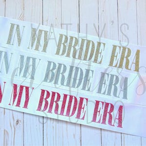 Puede incluir: Tres fajas de satén blanco con el texto "In My Bride Era" en purpurina dorada, plateada y rosa.