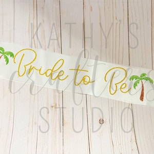 Peut inclure: Sash en satin blanc avec inscription dorée scintillante "Bride to Be" et deux motifs de palmiers verts.