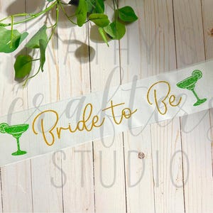May include: White satin sash with gold glitter lettering that says "Bride to Be" and two green glitter margarita glasses.