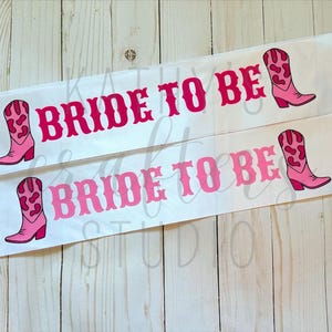 May include: Two white satin sashes with the text "BRIDE TO BE" in pink, accompanied by pink cowboy boot illustrations. The sashes are designed for a hen party or bridal shower.
