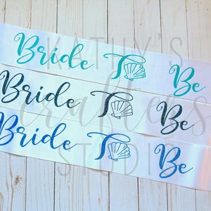 May include: Four white satin sashes with blue and teal glitter lettering. The sashes read "Bride to Be" and feature a seashell design.