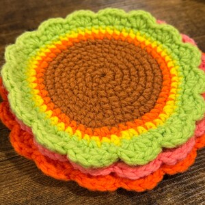 May include: A stack of colorful, handmade crochet coasters. The coasters feature a circular design with a scalloped edge. The colors include shades of orange, pink, yellow, green, and brown. The coasters are stacked on top of each other.