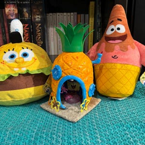 Può includere: Tre peluche del cartone animato SpongeBob. Un peluche giallo di SpongeBob a forma di Krabby Patty, un peluche rosa di Patrick Stella a forma di cono gelato e una casa a forma di ananas gialla con una porta blu.