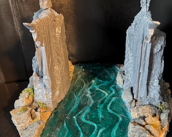 May include: A miniature diorama featuring two gray statues flanking a teal river. The statues, with outstretched arms, stand on rocky banks with patches of green moss. The base is a light brown, circular shape.