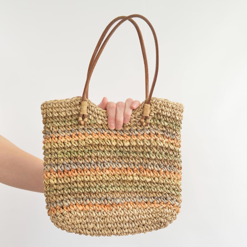 May include: A woven straw tote bag with brown leather handles. The bag features horizontal stripes of orange, green, and tan. The handles are attached with tan-colored woven accents.