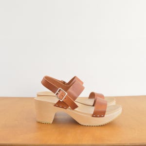 May include: Brown leather sandals with a wooden platform and heel. The sandals have two straps across the foot and ankle, secured with silver buckles. The wooden platform is approximately 3 inches high.