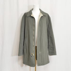 May include: A long, olive green jacket with a beige lining and a gold zipper. The jacket has a collar and is displayed on a mannequin. The jacket is a medium length and has pockets.