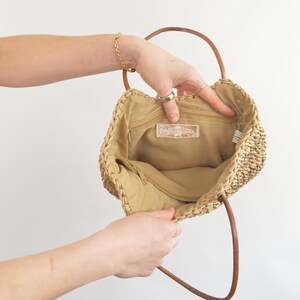 May include: Open view of a woven straw handbag with brown leather handles. The interior is tan with a zippered pocket and a label that reads "Cappelli". The bag is held open, showcasing the inside.