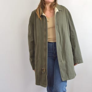 May include: Olive green jacket with a full-length zipper and a beige lining. The jacket has a collar and long sleeves. The model is wearing blue jeans and a beige top. The jacket appears to be made of a durable fabric.