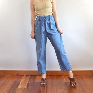 May include: Light blue denim pants with an elastic waistband and drawstring. The pants are paired with a beige tank top and brown leather clogs. The outfit is casual and comfortable, suitable for everyday wear.