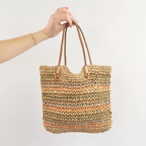 May include: A woven tote bag with brown leather handles. The bag features horizontal stripes of tan, orange, and green. The bag is held up by a person's hand, showing a gold bracelet and rings.