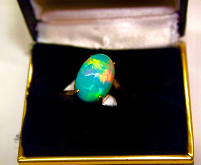 K&ouml;nnte beinhalten: Ein goldener Ring mit einem gro&szlig;en ovalen Opal-Edelstein. Der Opal hat eine leuchtende gr&uuml;ne, blaue und orange Farbe.