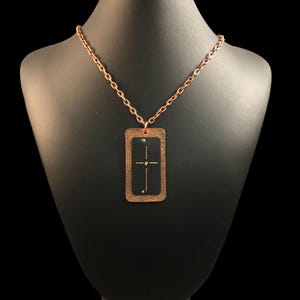 May include: A copper necklace with a rectangular pendant featuring a cross design with a small heart at the center. The pendant has a textured surface and a gold-colored outline.