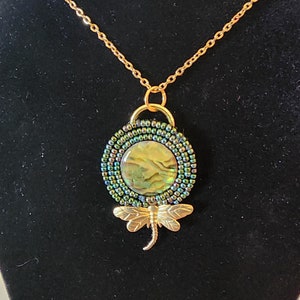 May include: A gold chain necklace with a green and iridescent abalone shell pendant surrounded by green beads. A gold dragonfly charm hangs below the pendant.