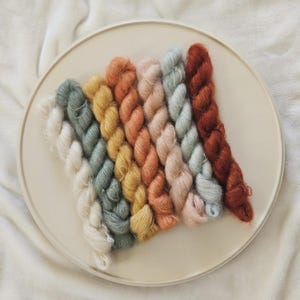May include: A selection of colourful yarn skeins arranged in a rainbow pattern on a cream-coloured embroidery hoop. The yarn colours include white, green, yellow, orange, pink, and brown. The yarn is soft and fluffy, ideal for knitting or crochet projects.