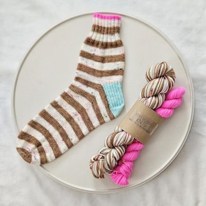 May include: A hand-knitted sock with brown and white stripes, a light blue heel and toe, and a pink cuff. Beside the sock are two skeins of yarn, one with brown and white variegated colours and the other hot pink. The yarn is labelled "Tiny Human Knits."