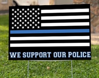 Pode incluir: Uma bandeira americana em preto e branco com uma faixa azul no meio. A bandeira está em um cartaz de jardim com o texto "WE SUPPORT OUR POLICE".