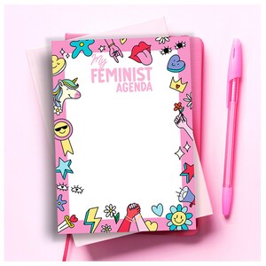 Puede incluir: Bloc de notas rosa con un interior blanco y un borde rosa. El borde está decorado con coloridas ilustraciones de dibujos animados que incluyen un unicornio, una estrella, una flor, un corazón, un rayo, un arcoíris y el texto "My Feminist Agenda".
