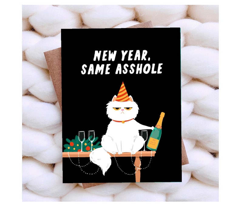 May include: A greeting card with a black background features a white cat wearing a party hat and holding a champagne bottle. The text reads "NEW YEAR, SAME ASSHOLE". Two champagne glasses and festive decorations are on the table.