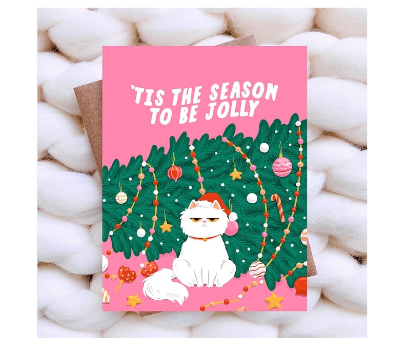 May include: A pink Christmas card with the text "'Tis the season to be jolly." The card features a grumpy white cat wearing a Santa hat, sitting in front of a decorated Christmas tree. The card is placed on a white, textured surface.