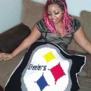 May include: A handmade, crocheted blanket featuring the Pittsburgh Steelers logo. The blanket has a black border, a white center with the team name, and the team's logo in yellow, red, and blue. The blanket is on a brown couch.