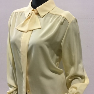 May include: A pale yellow long-sleeved blouse with a bow tie detail at the collar. The blouse has a button-up front and long sleeves with button cuffs.