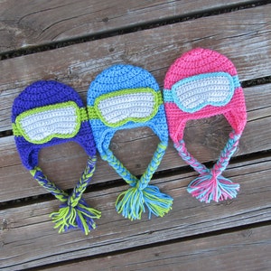 May include: Three crocheted hats with goggles and braided ties. The hats are blue, purple, and pink. The goggles are white with green trim. The ties are green, purple, and pink.