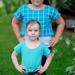 Puede incluir: Dos chicas con camisas azules. La chica mayor lleva una camisa a cuadros azules y blancos y la chica menor lleva una camisa azul lisa anudada a la cintura. Ambas chicas sonríen.